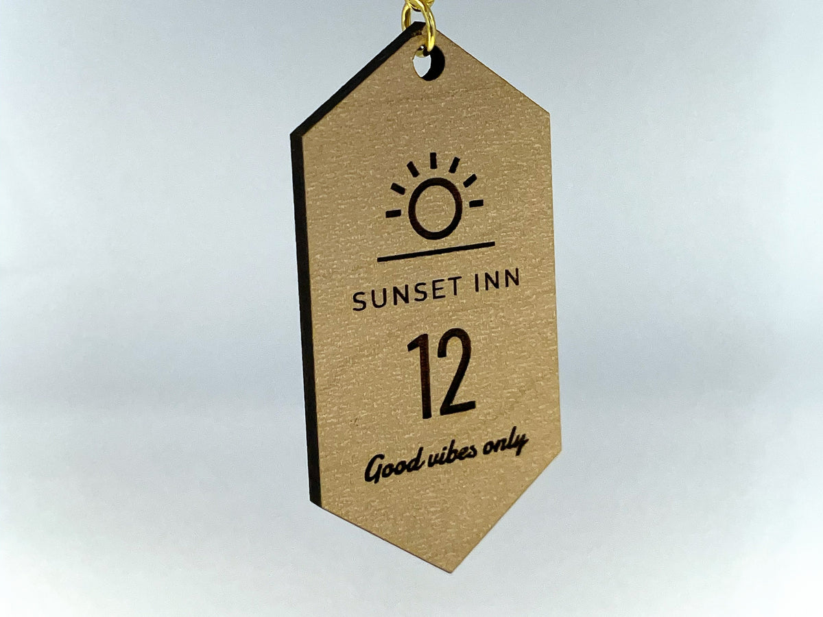 A motel tag keychain in maple finish hanging from its gold chain, engraved with a sun logo, SUNSET INN, the number 12 and Good vibes only.