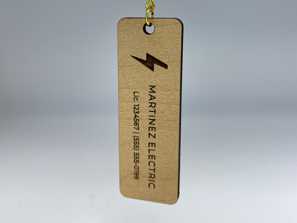A bar keychain in maple finish hanging from its gold chain, engraved with a lightning bolt logo, MARTINEZ ELECTRIC and a license and phone line.
