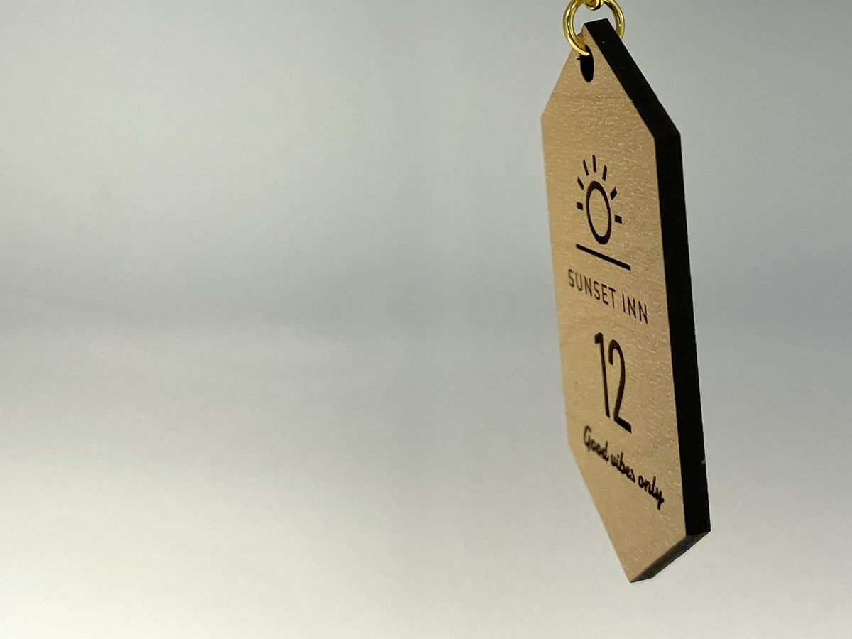 The motel tag keychain seen nearly edge on, showing the thickness of the board and its dark cut edge, on a gold chain.