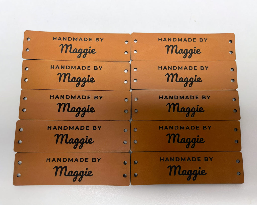Ten rawhide leatherette knitting labels in two rows, each engraved HANDMADE BY over the name Maggie in script, with two sewing holes at each end.