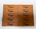 Ten rawhide leatherette knitting labels in two rows, each engraved HANDMADE BY over the name Maggie in script, with two sewing holes at each end.