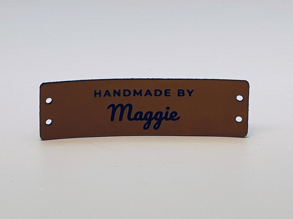 Rawhide leatherette knitting label standing on a table, engraved HANDMADE BY over the name Maggie in script, with two sewing holes at each end.