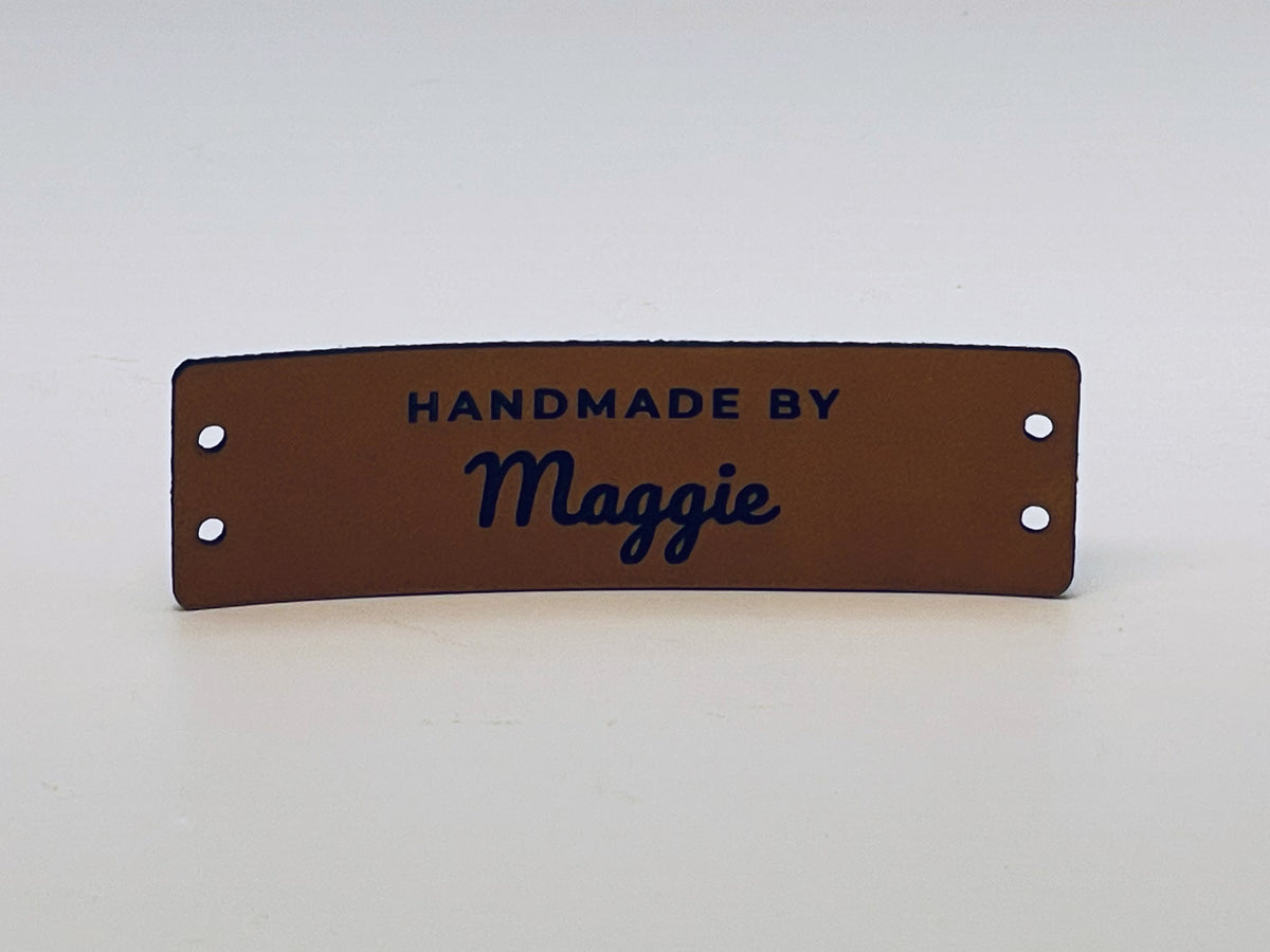 Rawhide leatherette knitting label standing on a table, engraved HANDMADE BY over the name Maggie in script, with two sewing holes at each end.