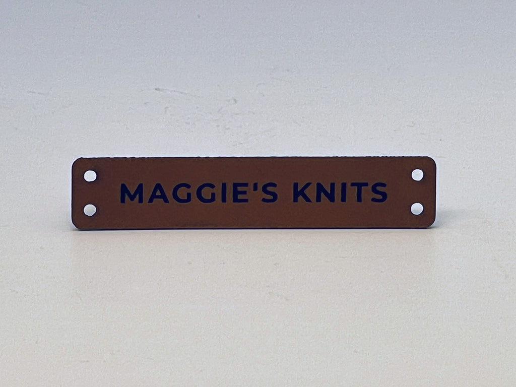 Slim rawhide leatherette label engraved MAGGIE'S KNITS in bold capitals, with two sewing holes at each end.