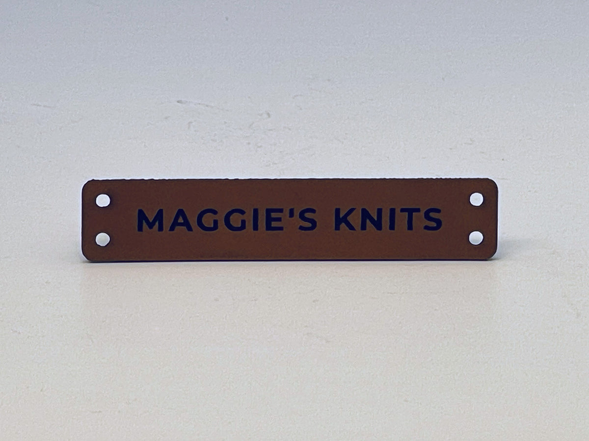 Slim rawhide leatherette label engraved MAGGIE'S KNITS in bold capitals, with two sewing holes at each end.