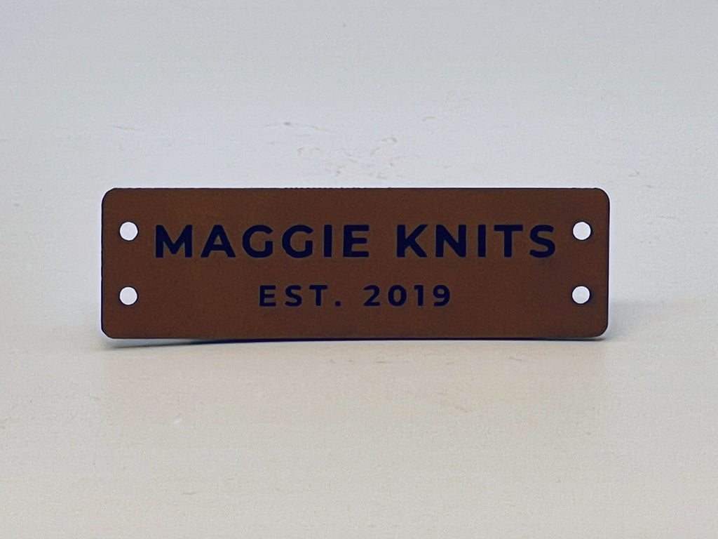 Rawhide leatherette label engraved MAGGIE KNITS over EST. 2019, with two sewing holes at each end.