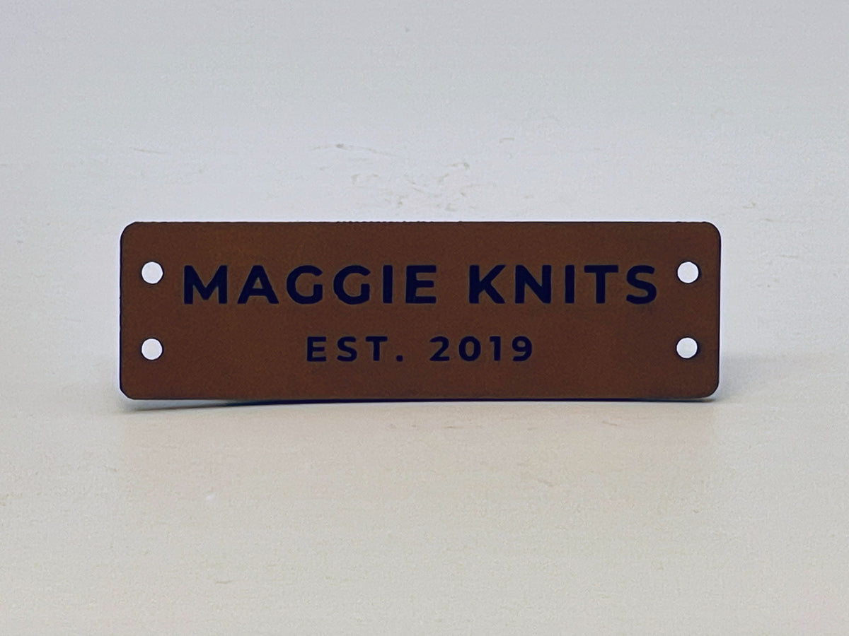 Rawhide leatherette label engraved MAGGIE KNITS over EST. 2019, with two sewing holes at each end.