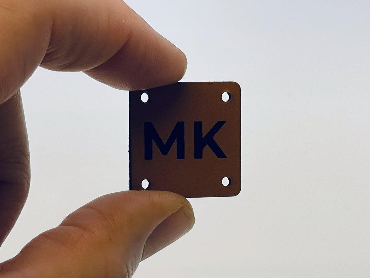A hand holding a small square rawhide leatherette label engraved with the monogram MK, with a sewing hole in each corner.