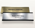 Two rectangular desk nameplates with black engraved text. The top plate is gold-toned and metal-look, with the name VERONICA SANDOVAL and the title Board President. The bottom plate is silver-toned and metal-look, with the name SARAH MITCHELL and the title Store Manager.