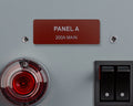 A rectangular dark red identification label with white text reading PANEL A and 200A MAIN, mounted on a light gray surface between two silver screws, positioned above a circular red indicator light and a black dual rocker switch.