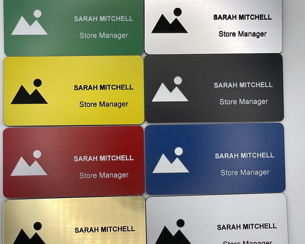 A collection of rectangular name badges in green, yellow, red, gold, silver, black, and blue. Each badge features a mountain and sun icon, the name SARAH MITCHELL, and the title Store Manager engraved in black or white text.