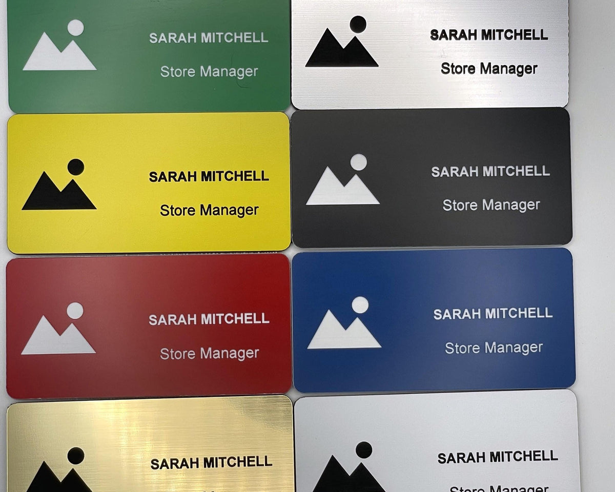 A collection of rectangular name badges in green, yellow, red, gold, silver, black, and blue. Each badge features a mountain and sun icon, the name SARAH MITCHELL, and the title Store Manager engraved in black or white text.