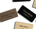 Rectangular name badges in black, light wood, and dark wood finishes. The dark wood badge features the engraved text Willow Grant Herbalist. The black badge displays the engraved text COLE BRAM Resident DJ.