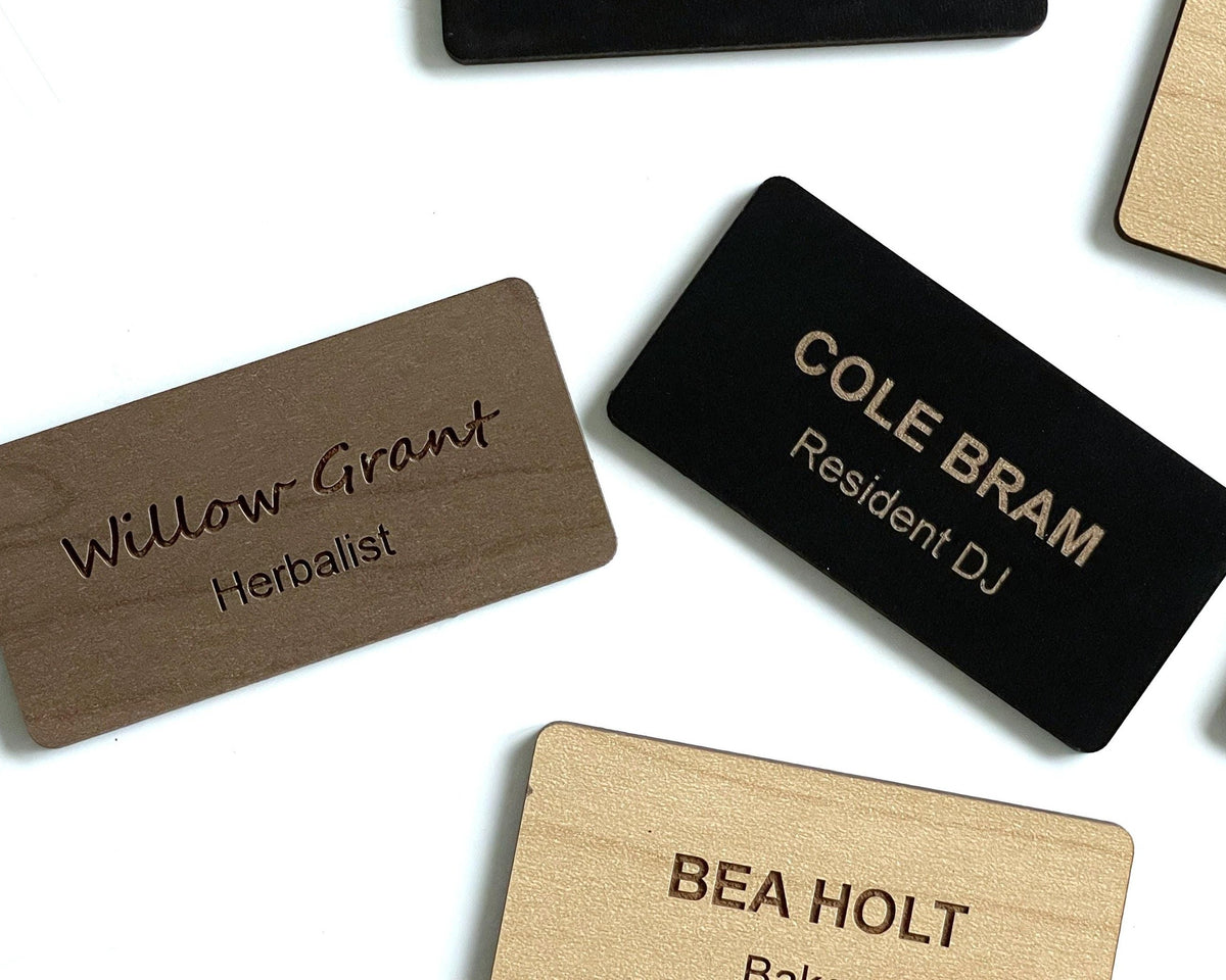 Rectangular name badges in black, light wood, and dark wood finishes. The dark wood badge features the engraved text Willow Grant Herbalist. The black badge displays the engraved text COLE BRAM Resident DJ.