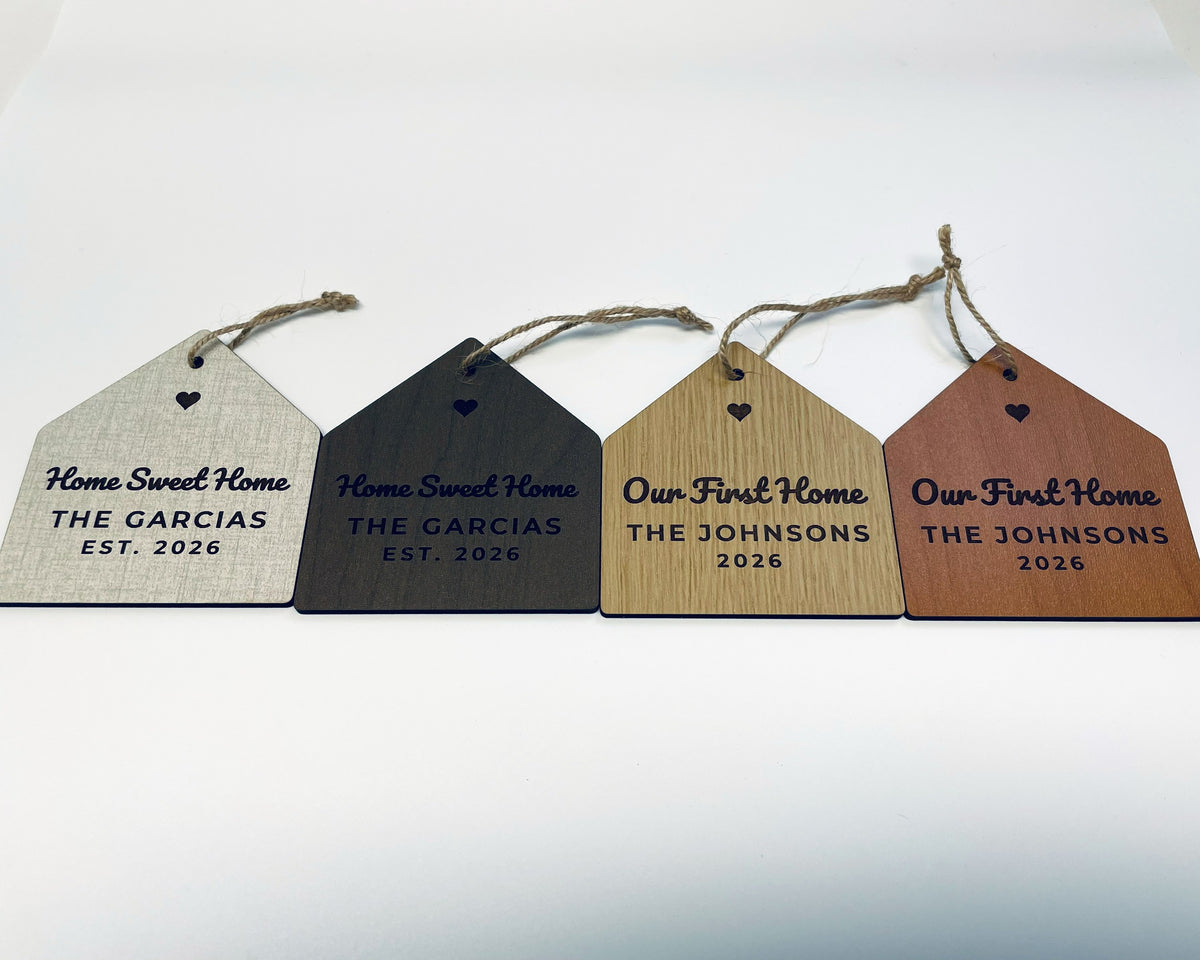 Four house-shaped Christmas ornaments lying in a row on jute twine, each with a cut-out heart: Home Sweet Home, THE GARCIAS, EST. 2026 in linen and walnut finishes, and Our First Home, THE JOHNSONS, 2026 in oak and cherry finishes.