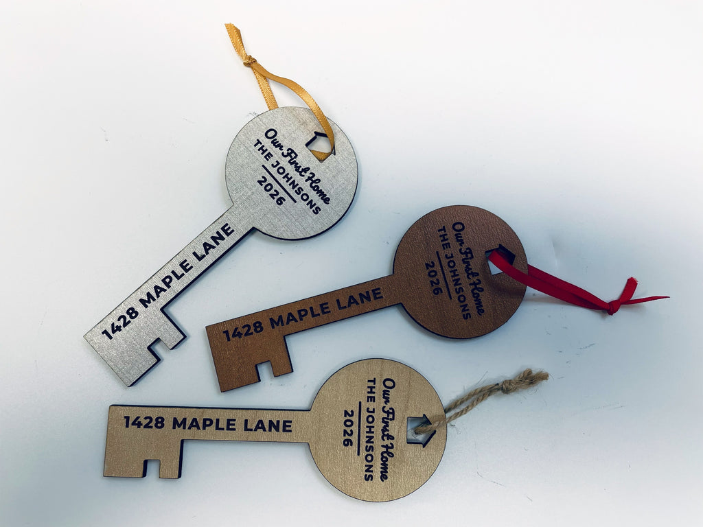 Three key-shaped first home ornaments lying flat in linen, cherry and oak finishes, each with a little house cut out of the round top for the ribbon: Our First Home, THE JOHNSONS, 2026 in the top and 1428 MAPLE LANE down the shaft.