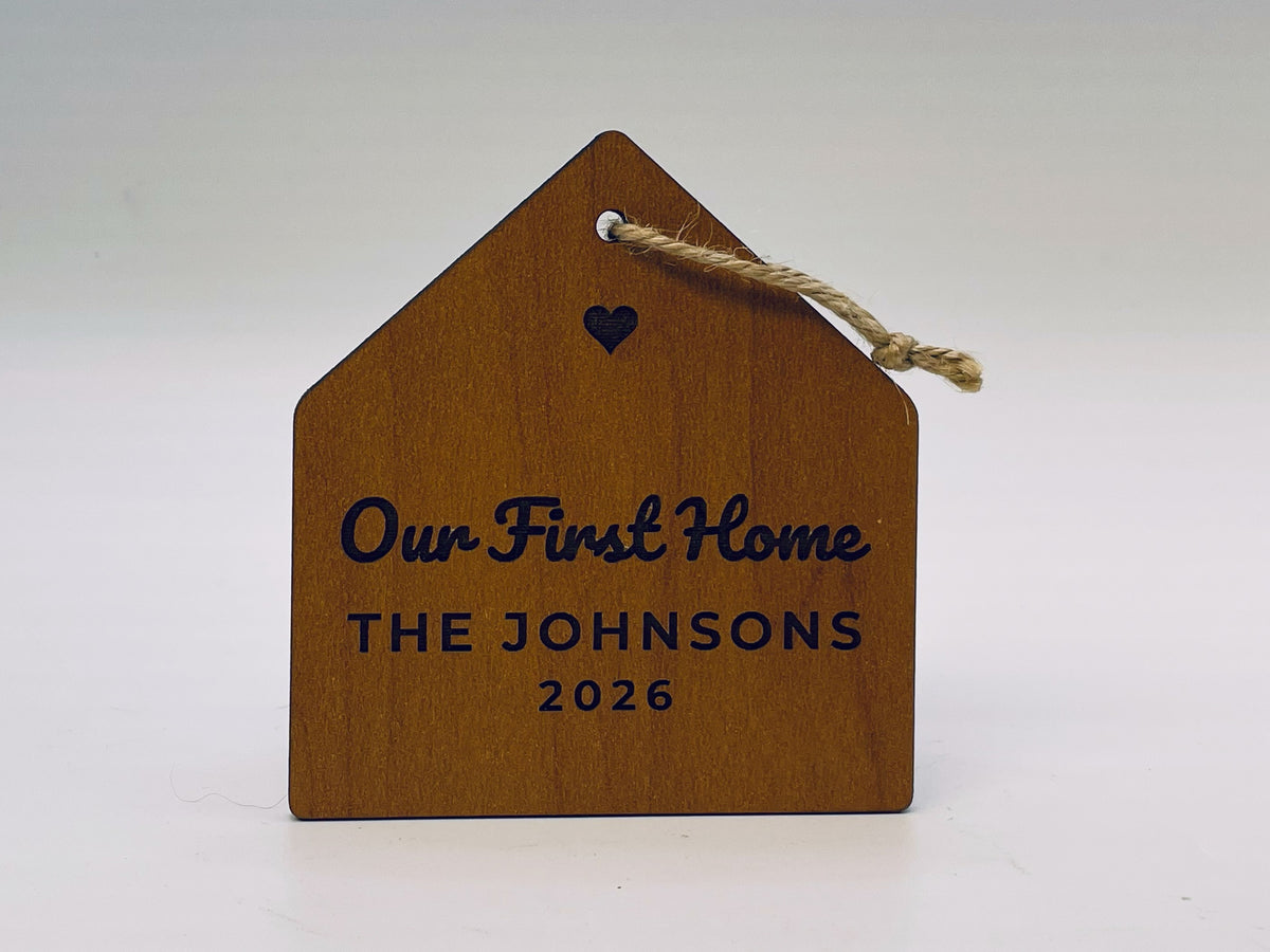 House-shaped ornament in a cherry finish on jute twine: a cut-out heart, Our First Home in script, THE JOHNSONS and 2026.