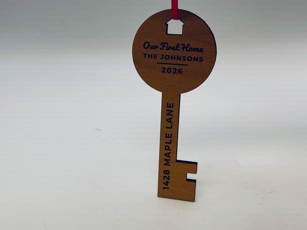 The key ornament in a cherry finish standing upright on red ribbon: Our First Home, THE JOHNSONS and 2026 in the round top and 1428 MAPLE LANE down the shaft.