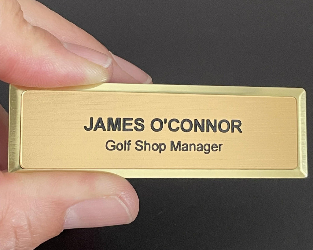 A name badge on a gold metal bar with a brushed metal-look face. The text reads JAMES O'CONNOR and Golf Shop Manager in black engraved lettering. The badge has a clean, professional appearance.