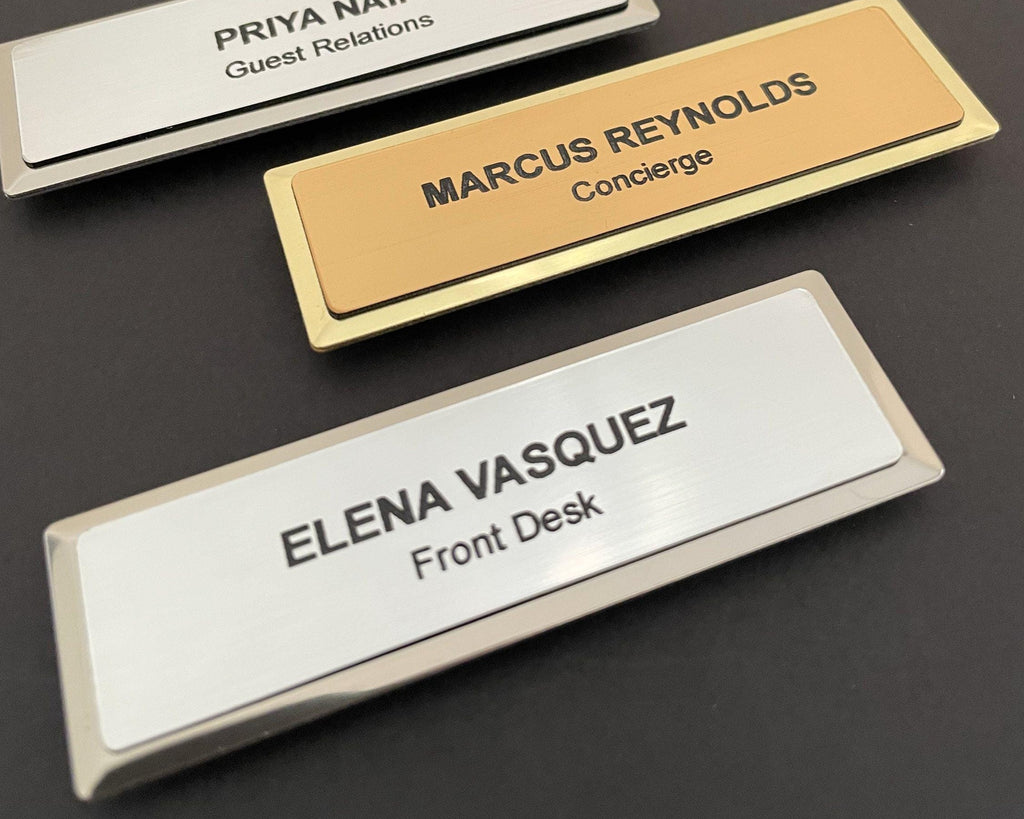 Three name badges on metal bars. One gold badge reads MARCUS REYNOLDS Concierge. Two silver badges read PRIYA NAIR Guest Relations and ELENA VASQUEZ Front Desk.
