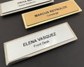 Three name badges on metal bars. One gold badge reads MARCUS REYNOLDS Concierge. Two silver badges read PRIYA NAIR Guest Relations and ELENA VASQUEZ Front Desk.