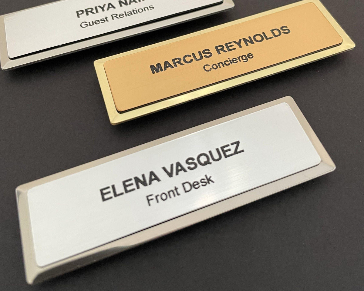 Three name badges on metal bars. One gold badge reads MARCUS REYNOLDS Concierge. Two silver badges read PRIYA NAIR Guest Relations and ELENA VASQUEZ Front Desk.