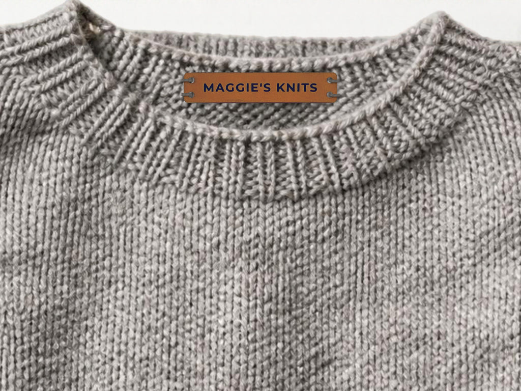 Rawhide leatherette label engraved MAGGIE'S KNITS, sewn with gray yarn inside the back collar of a gray knitted sweater.