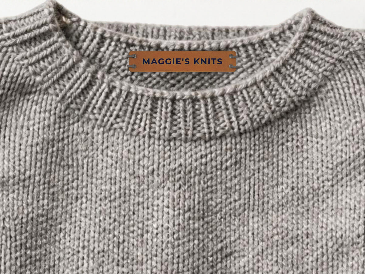 Rawhide leatherette label engraved MAGGIE'S KNITS, sewn with gray yarn inside the back collar of a gray knitted sweater.