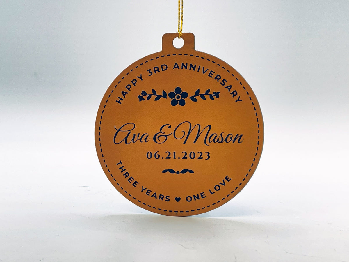 Rawhide leatherette Christmas ornament, a round with a cap on top, hanging from gold string: HAPPY 3RD ANNIVERSARY arched over a blossom and sprigs, Ava & Mason in script, 06.21.2023, and THREE YEARS, a heart, ONE LOVE along the bottom, inside a stitched border.