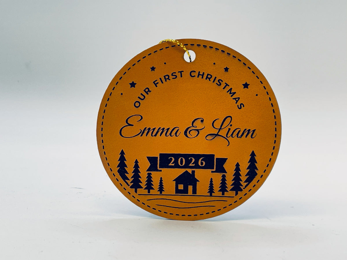 Round rawhide leatherette ornament: OUR FIRST CHRISTMAS arched with stars, Emma & Liam in script, 2026 on a ribbon banner, and a pine forest with a little cabin, inside a stitched border.