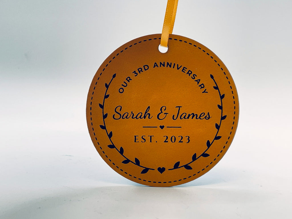 Round rawhide leatherette ornament on gold ribbon: OUR 3RD ANNIVERSARY arched over Sarah & James in script, a heart rule and EST. 2023, inside a laurel wreath and a stitched border.