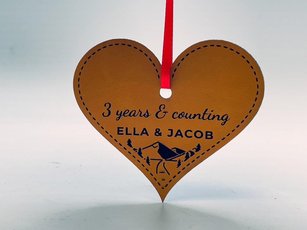 Heart-shaped rawhide leatherette ornament on red ribbon: 3 years & counting in script, ELLA & JACOB, and a mountain, pine and river scene in the point, inside a stitched border.