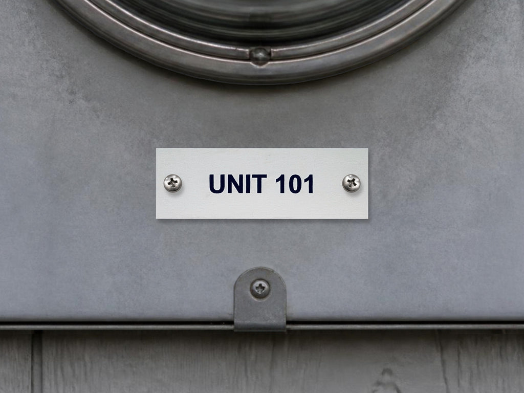 White engraved UNIT 101 label with black letters, screwed to a gray electric meter socket cover under the meter.