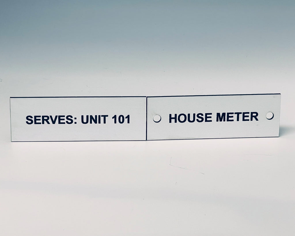 Two white engraved labels side by side with screw holes, black letters reading SERVES: UNIT 101 and HOUSE METER.