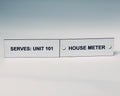 Two white engraved labels side by side with screw holes, black letters reading SERVES: UNIT 101 and HOUSE METER.