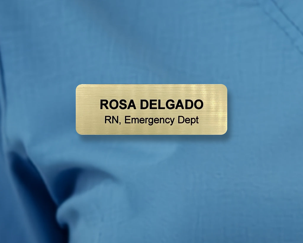 A rectangular gold-toned name badge with a brushed metal-look finish. The text reads ROSA DELGADO, RN, Emergency Dept in black sans-serif font.