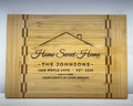Personalized bamboo cutting board with a butcher block inlay stripe near each end, engraved with a roof line and chimney, Home Sweet Home in script, THE JOHNSONS, 1428 MAPLE LANE, EST. 2026, and COMPLIMENTS OF AVERY BROOKS: the Home Sweet Home design on the medium board.