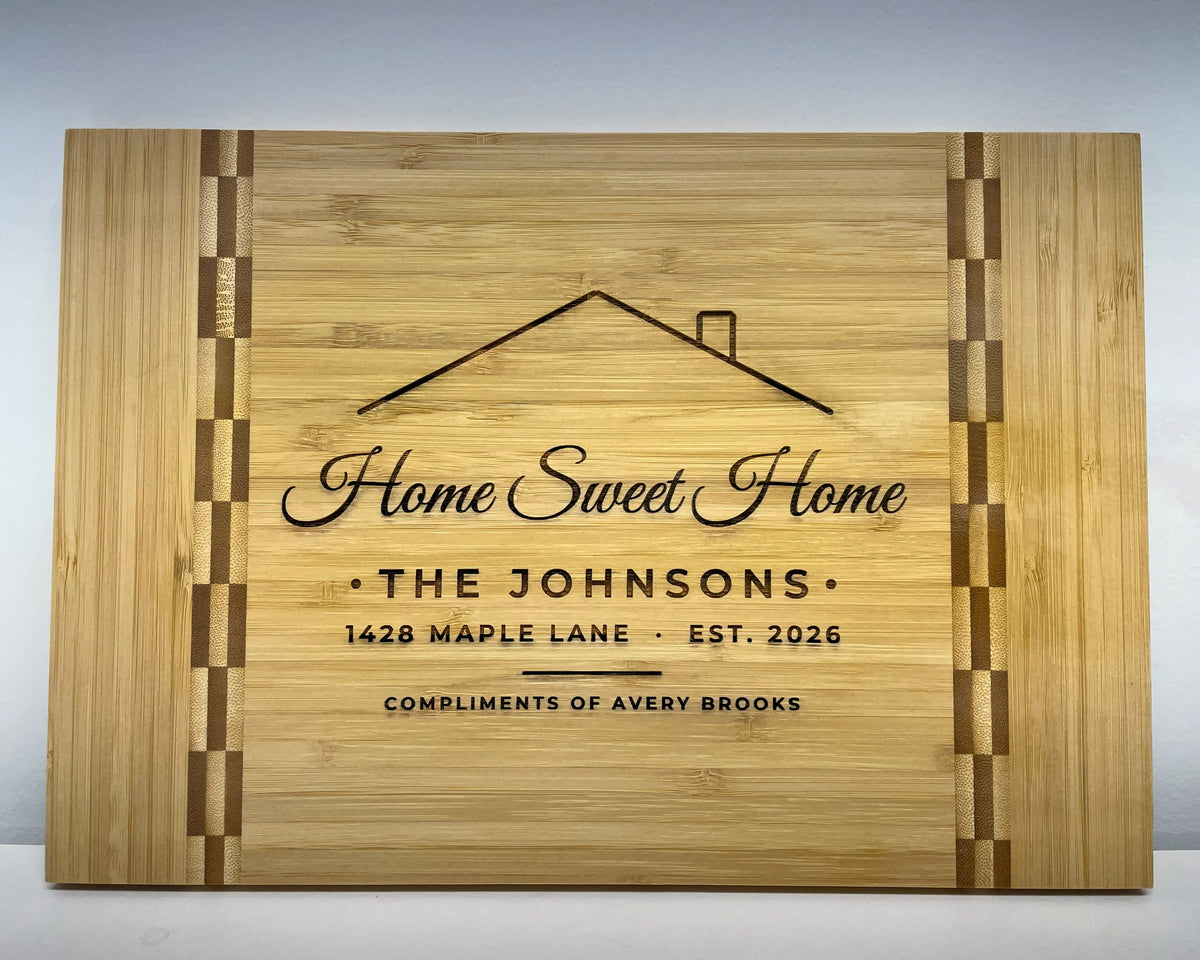 Personalized bamboo cutting board with a butcher block inlay stripe near each end, engraved with a roof line and chimney, Home Sweet Home in script, THE JOHNSONS, 1428 MAPLE LANE, EST. 2026, and COMPLIMENTS OF AVERY BROOKS: the Home Sweet Home design on the medium board.