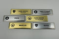 Six premium name badges on gold and silver metal bars, each engraved with a company logo at the left and a name, some with a title.