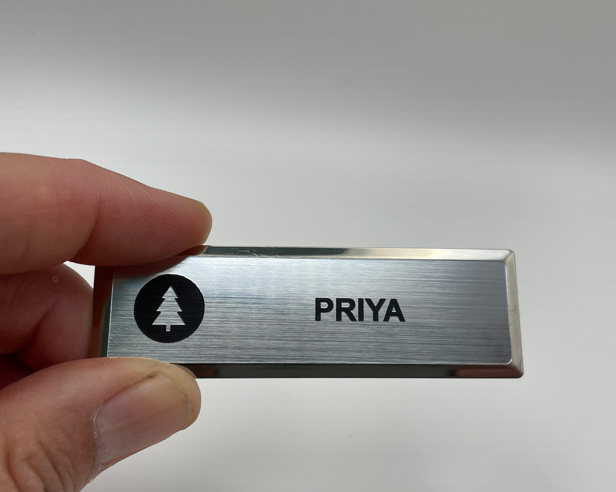 A silver metal bar name badge held between two fingers, engraved with a round black tree logo and the first name PRIYA.