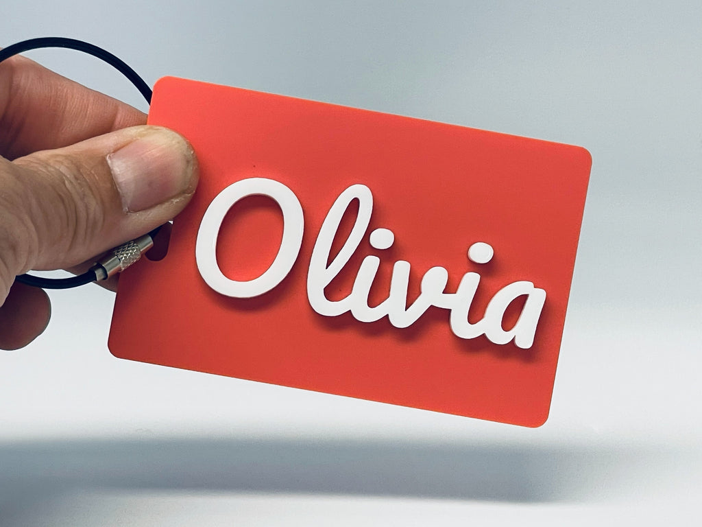A hand holding a coral rectangle bag tag with Olivia in raised white script letters.