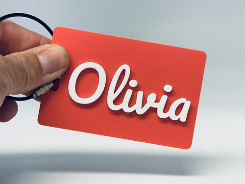 A hand holding a coral rectangle bag tag with Olivia in raised white script letters.