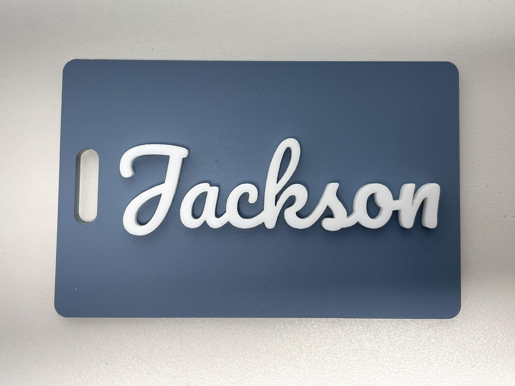 Slate blue rectangle bag tag standing on a table, with the name Jackson in raised white script letters.