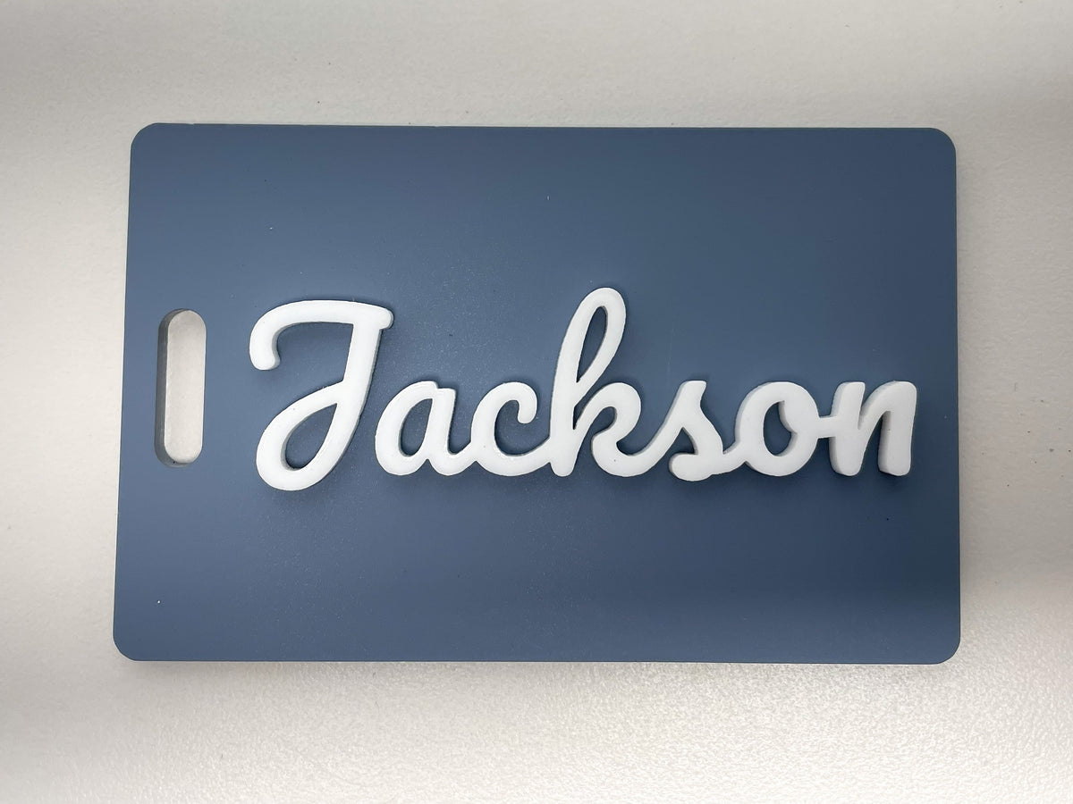 Slate blue rectangle bag tag standing on a table, with the name Jackson in raised white script letters.