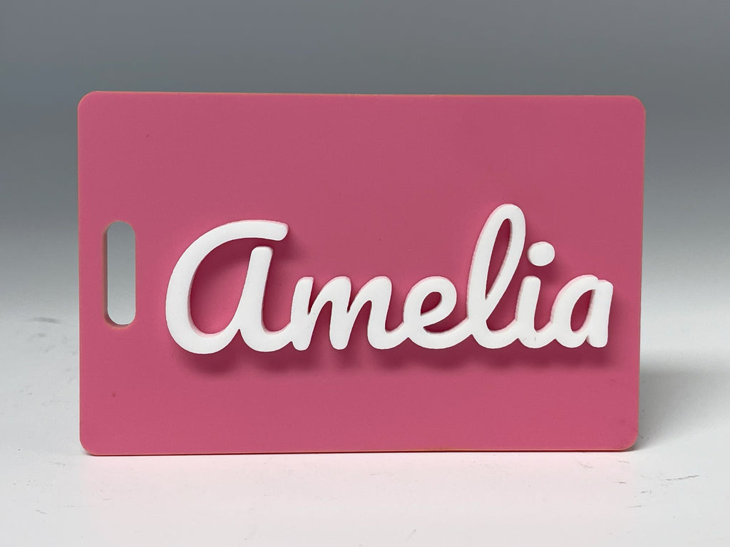Bubblegum pink rectangle bag tag standing on a table, with the name Amelia in raised white script letters.