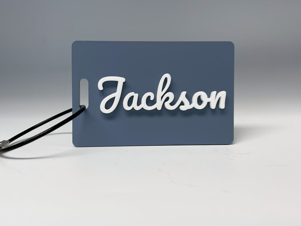The slate blue Jackson bag tag standing with its black wire loop, the name in raised white script letters.