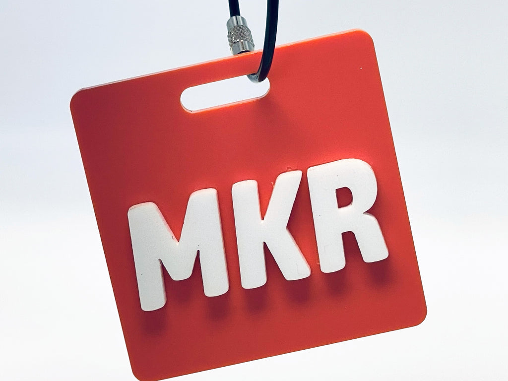 Square coral bag tag hanging from its wire loop, with the initials MKR in raised white letters.