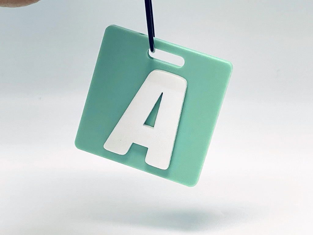 Square sage green bag tag hanging from its wire loop, with the initial A in a raised white letter.