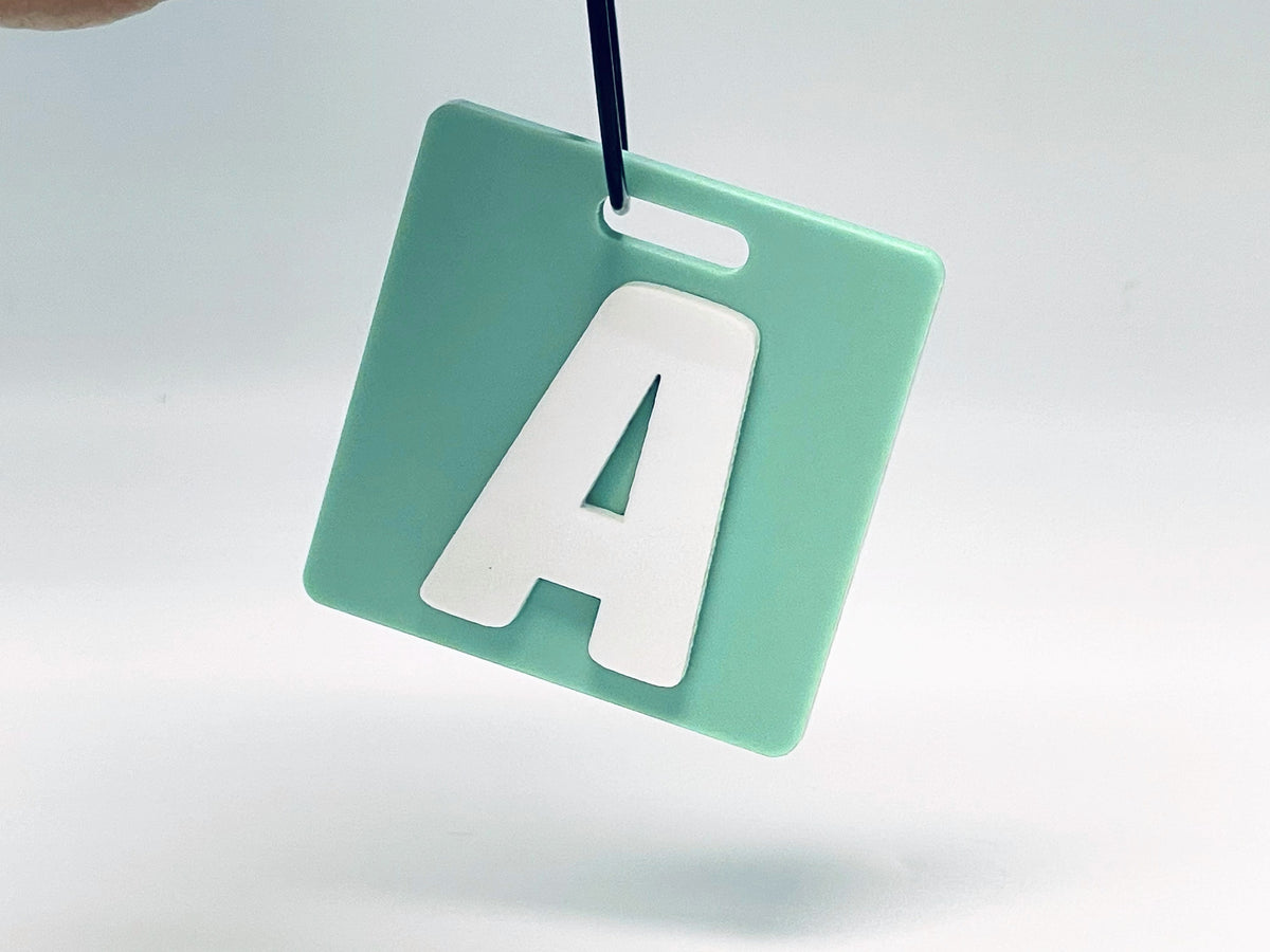 Square sage green bag tag hanging from its wire loop, with the initial A in a raised white letter.