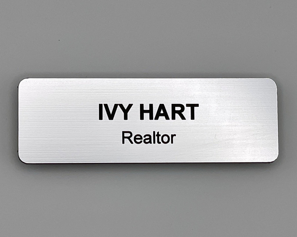 A rectangular silver name badge with a brushed metal-look finish. The text IVY HART is engraved in bold black capital letters on the top line, with the word Realtor in smaller black letters below.